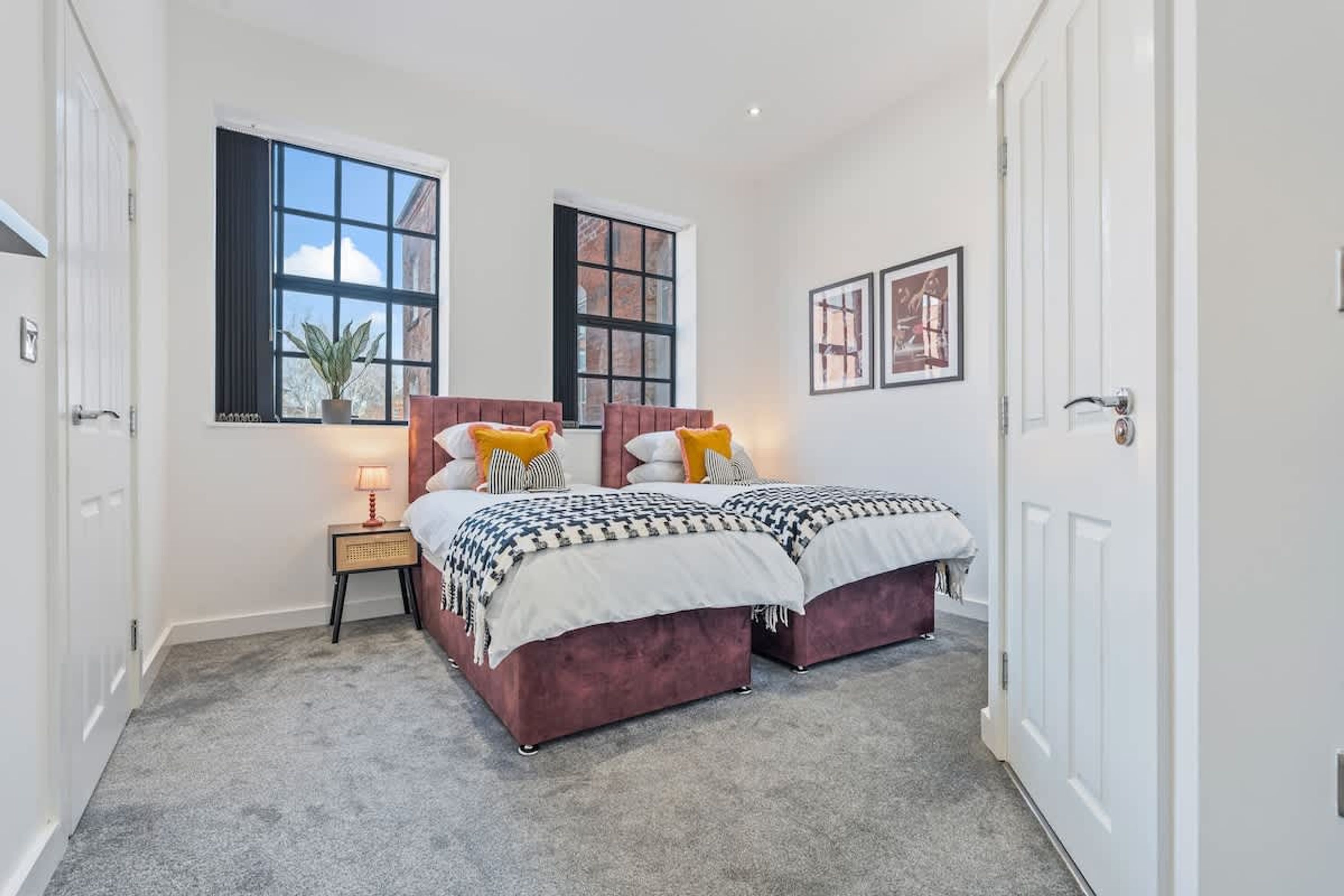 A bedroom with two beds, a nightstand, a lamp, and two windows.