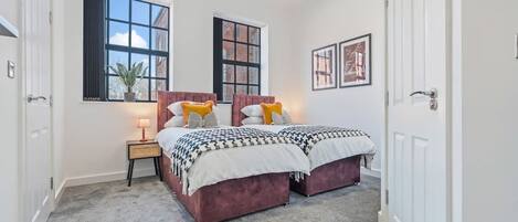 A bedroom with two beds, a nightstand, a lamp, and two windows.