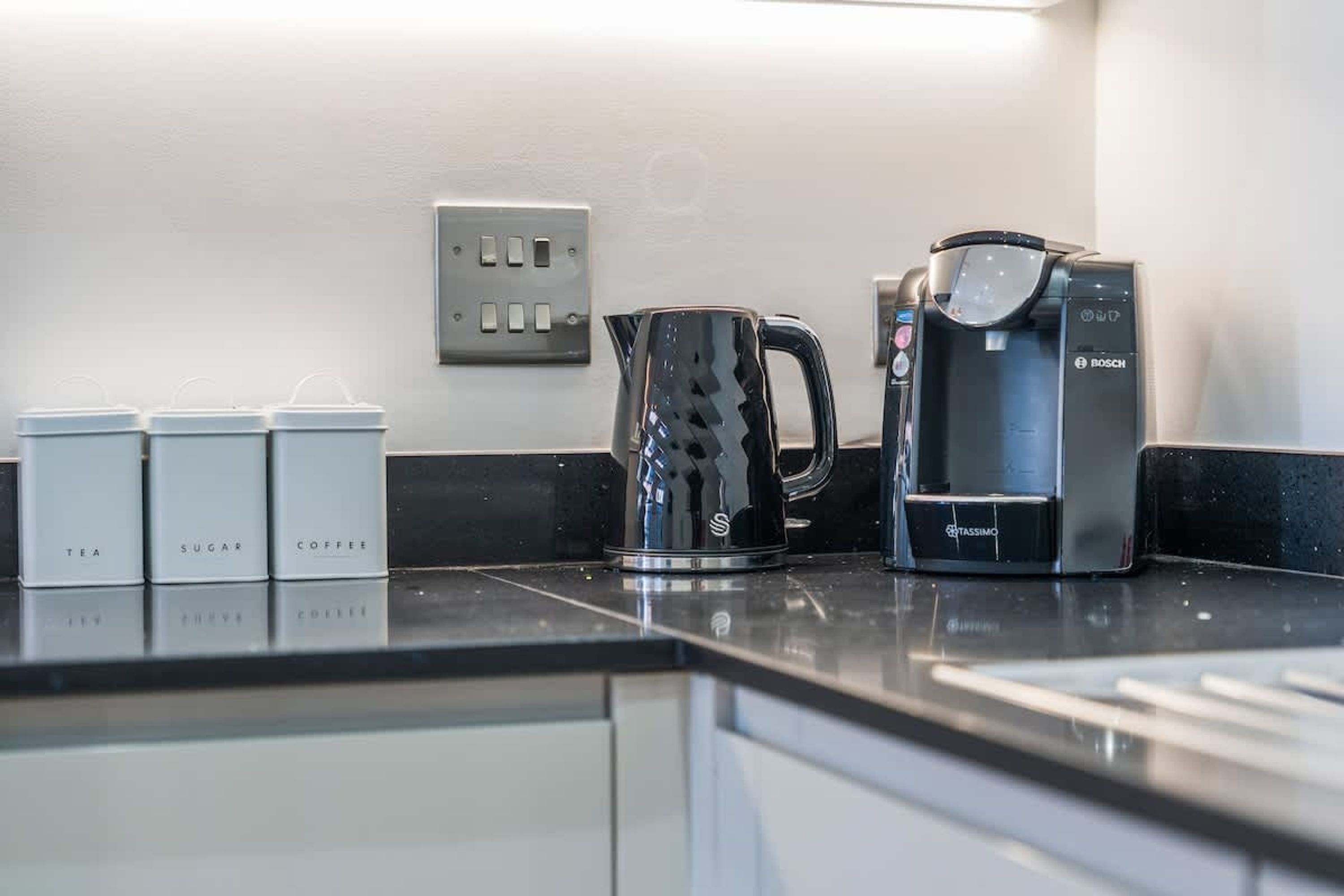 Eine Küchenarbeitsplatte mit Kaffeemaschine, Wasserkocher und beschrifteten Behältern für Tee, Zucker und Kaffee.
