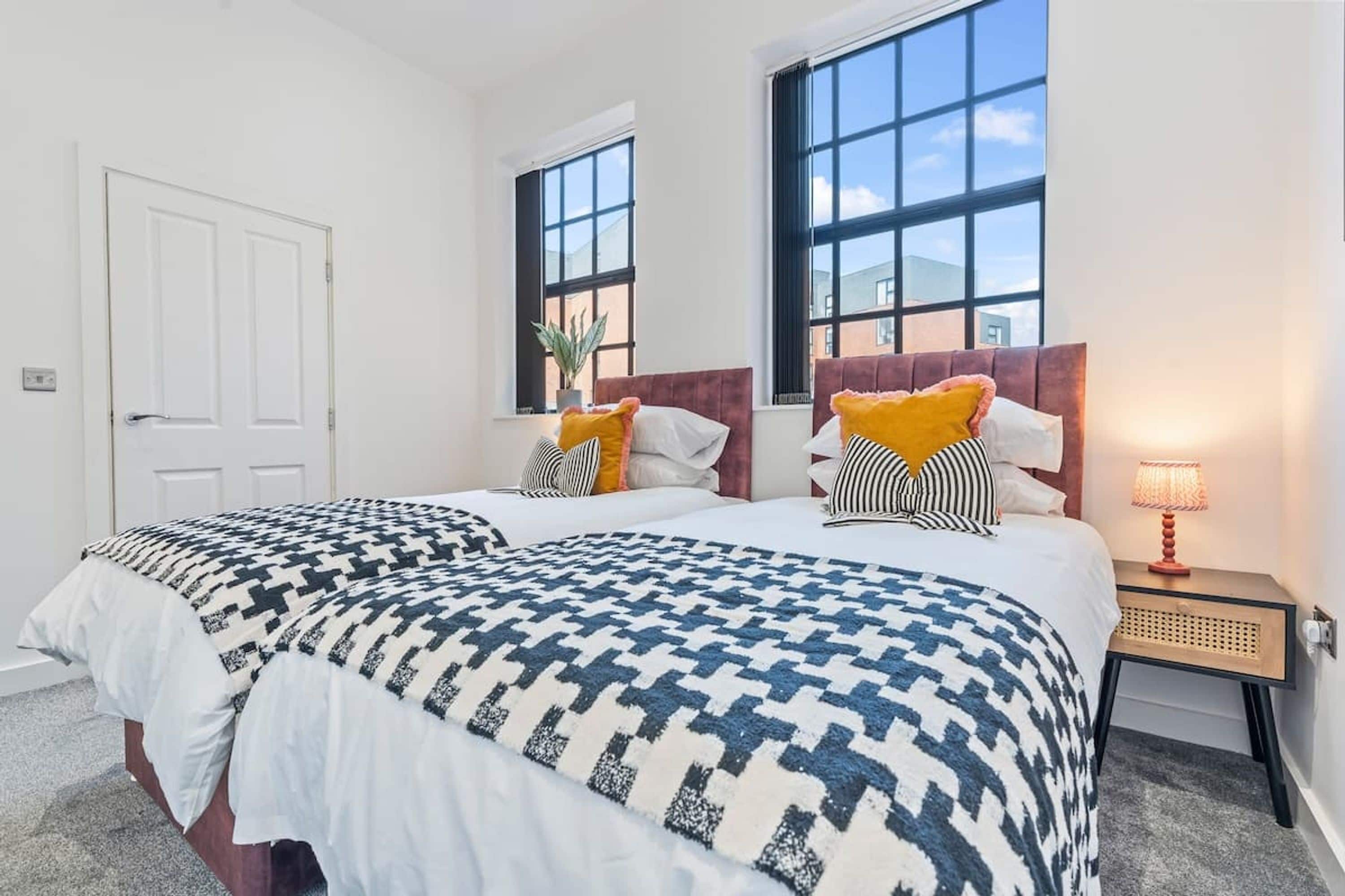 Quarto de hotel com duas camas, janelas amplas, mesa de cabeceira com abajur e uma porta.