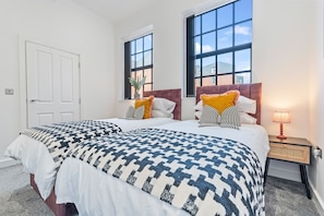 Quarto de hotel com duas camas, janelas amplas, mesa de cabeceira com abajur e uma porta.