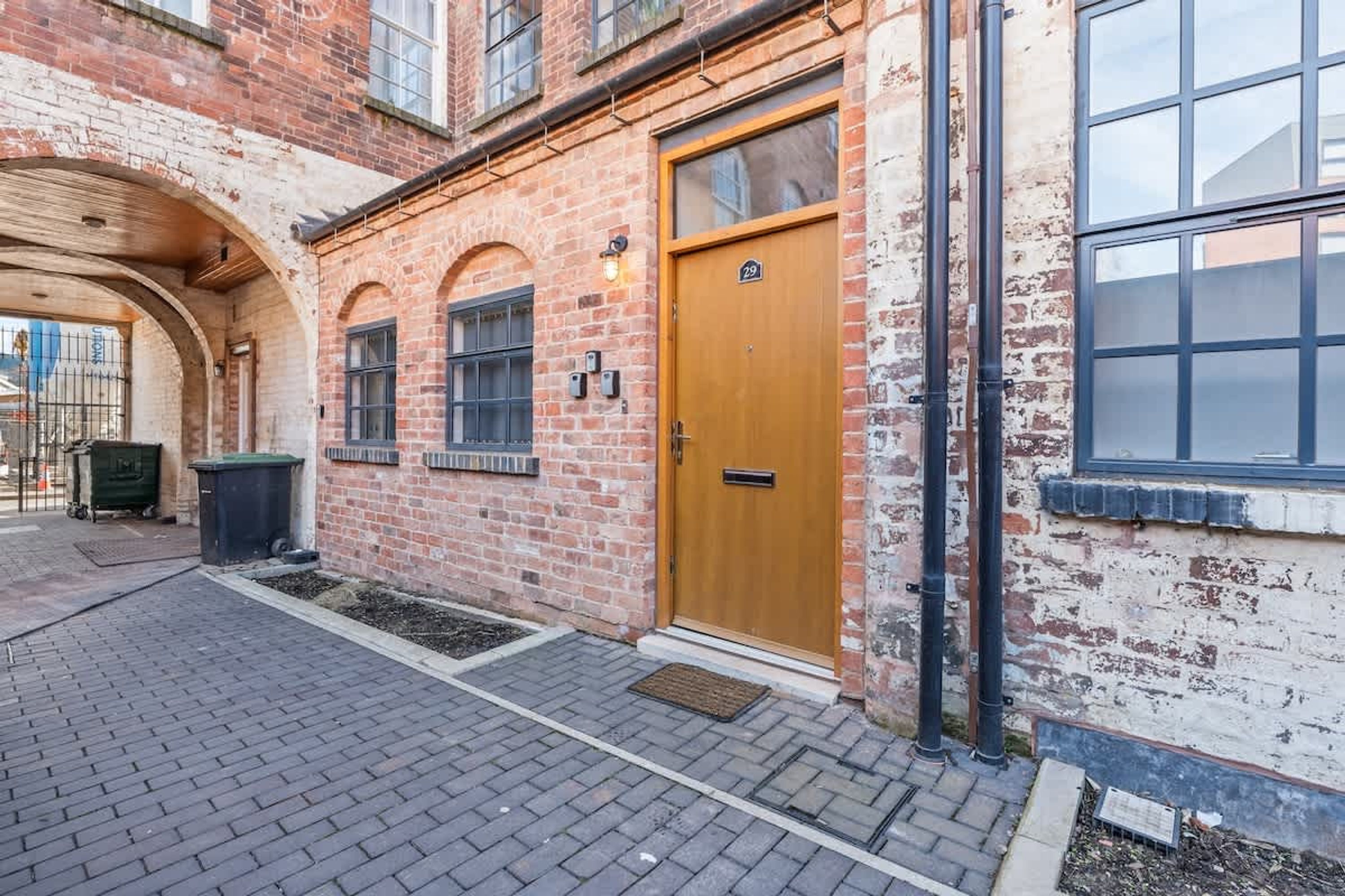 Een gebouw van baksteen met een houten deur, ramen en een klein buitenruimte met een verhard voetpad.