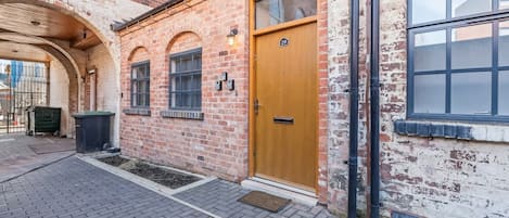 Un bâtiment en briques avec une porte en bois, des fenêtres et un petit espace extérieur doté d’un chemin pavé.