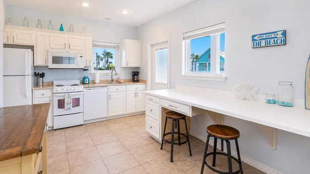 A kitchen with white cabinets, a stove, a microwave, and a window with a view of a blue building. - Pier Pressure Pools Beach Access Sunset View (Port Aransas)