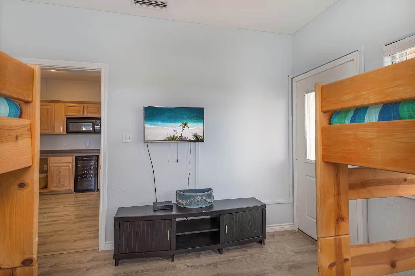 A room with bunk beds, a TV on the wall, a small cabinet, and a kitchen area in the background.