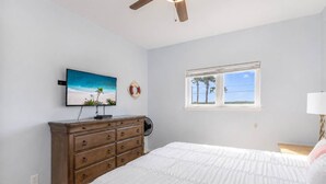 A bedroom with a bed, a dresser, a ceiling fan, a TV, a window with a view, and a wall-mounted life preserver. - Pier Pressure Pools Beach Access Sunset View (Port Aransas)
