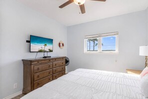 A bedroom with a bed, a dresser, a ceiling fan, a TV, a window with a view, and a wall-mounted life preserver. - Pier Pressure Pools Beach Access Sunset View (Port Aransas)