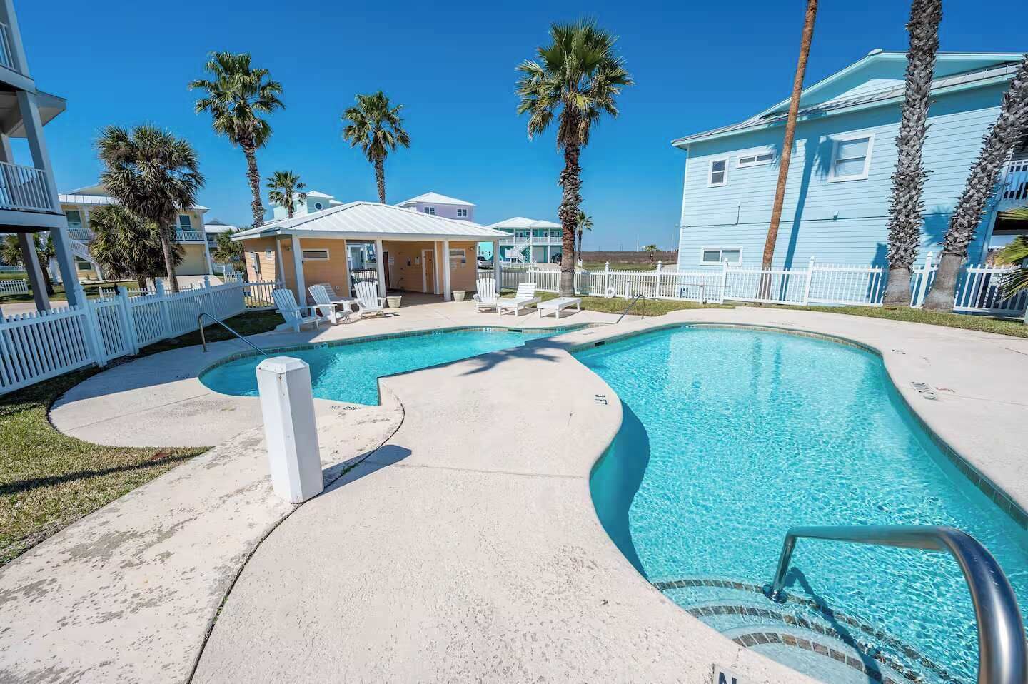 A swimming pool with lounge chairs, palm trees, and a white fence.