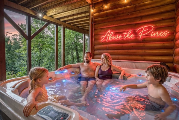 A hot tub with a neon sign reading 'Above the Pines' and a view of trees through large windows.