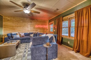 A cozy cabin interior with a wooden ceiling, a ceiling fan, a sectional sofa, a small table, and a window with curtains.