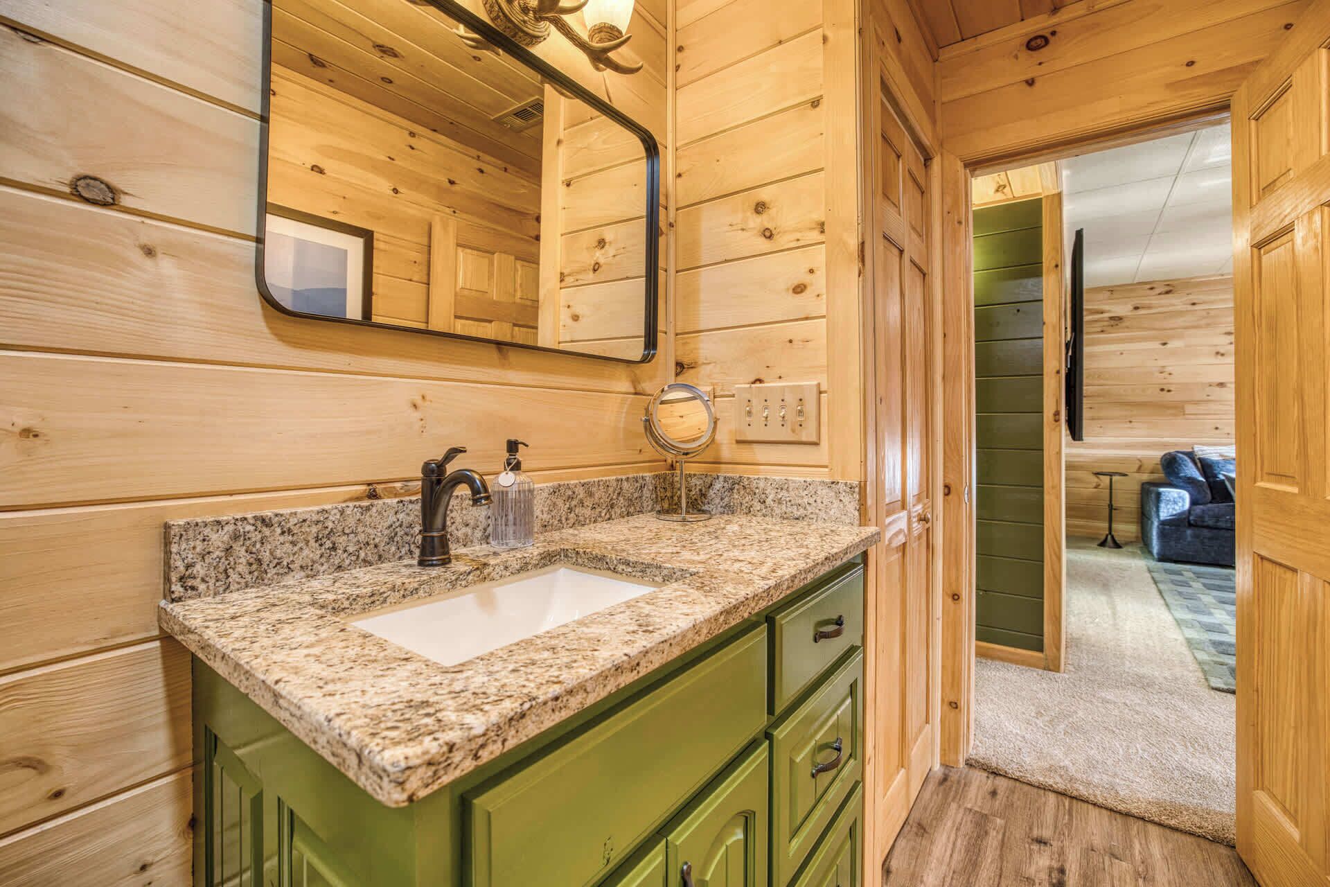 L’intérieur d’une cabane en bois est doté d’un évier avec un plan de travail en granit, de meubles de rangement verts, d’un miroir et d’une porte donnant sur une autre pièce.