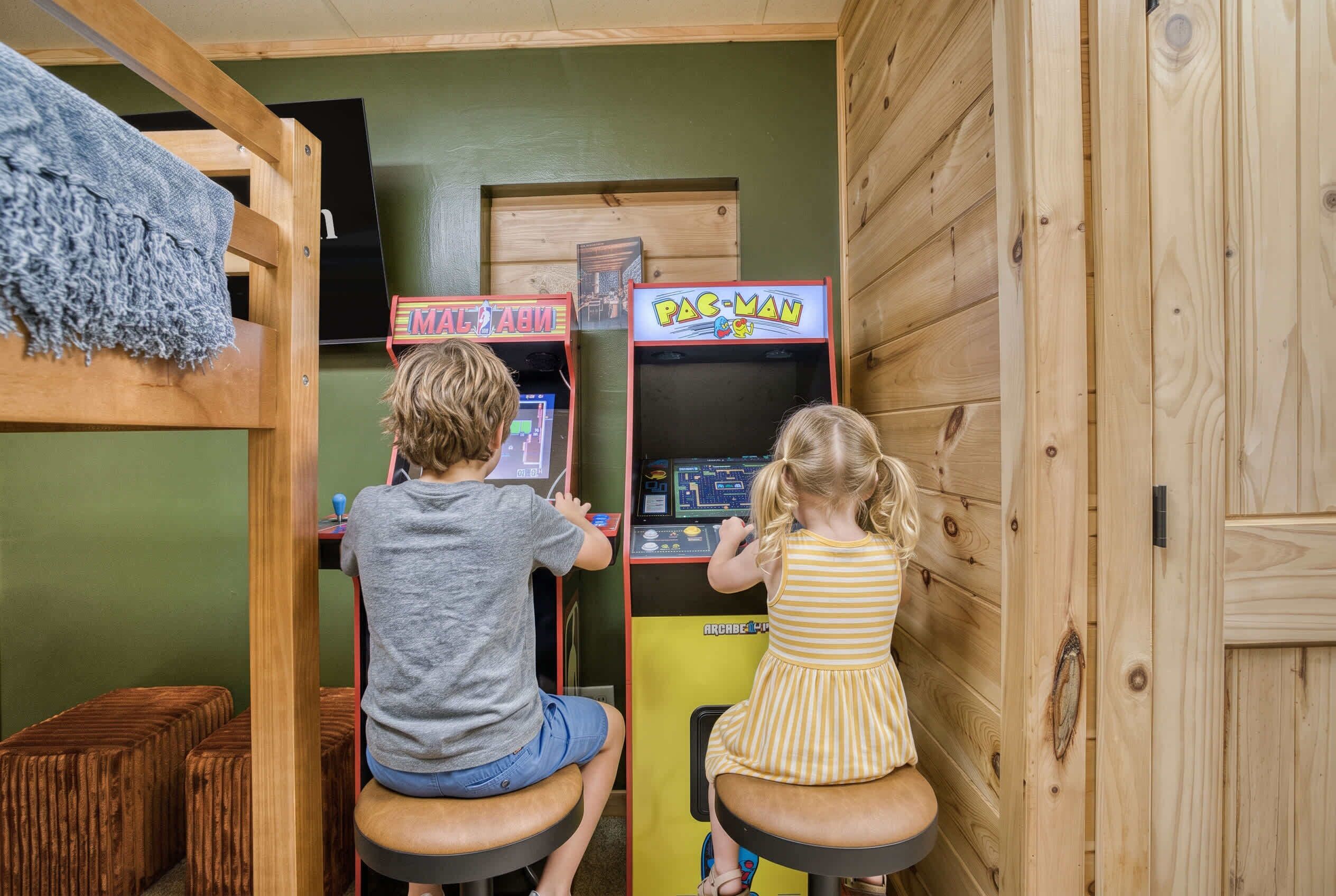 Two children playing arcade games in a room with wooden walls and a bunk bed.