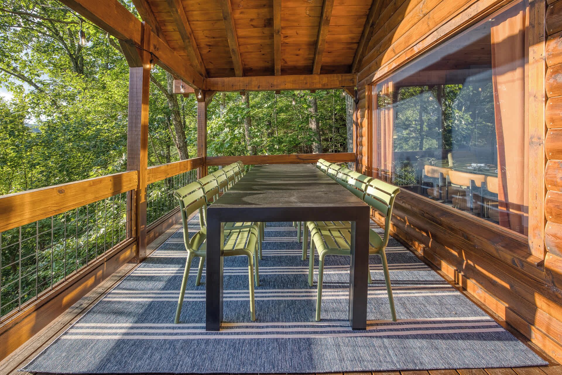 A covered outdoor seating area with a long table and chairs, surrounded by glass walls and wooden railings, overlooking a forested area.