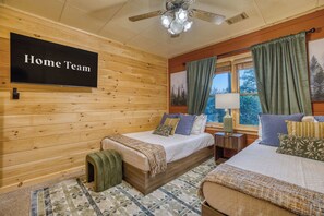 A room with two beds, a ceiling fan, a mounted TV, and a window with curtains.