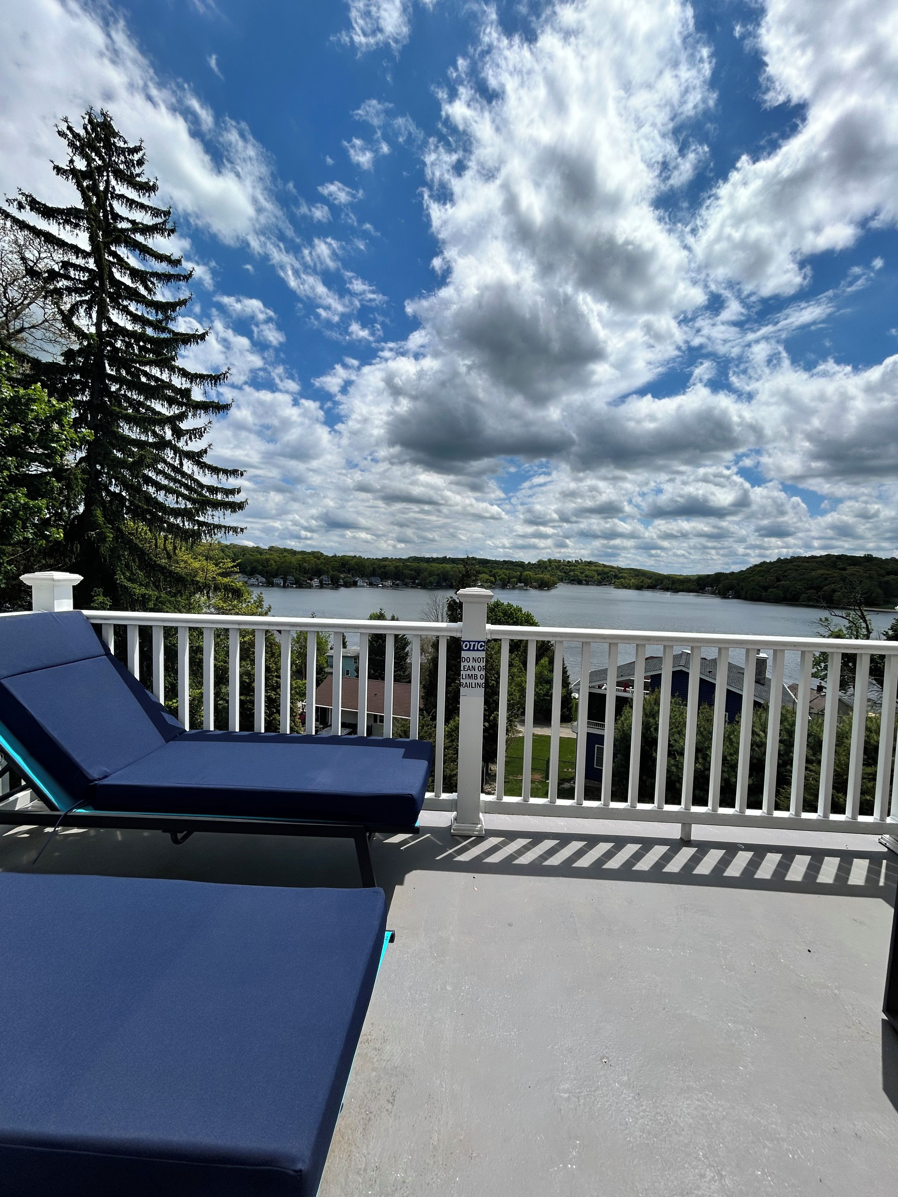 A balcony with a view of a lake and distant hills, a white railing, and a sign warning against leaning on it.