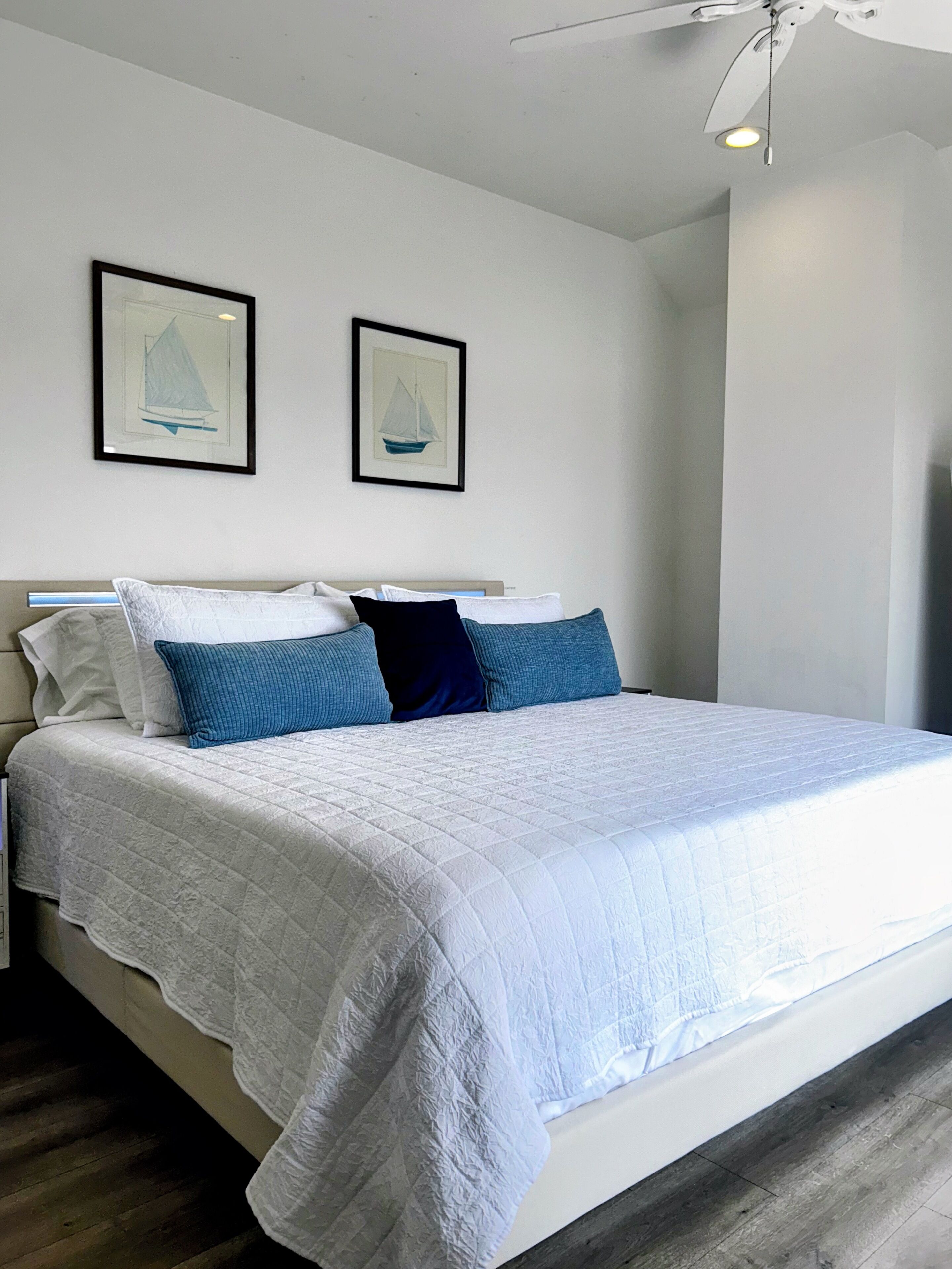 A neatly made bed with a white quilt and blue pillows, a ceiling fan, and two framed pictures on the wall.
