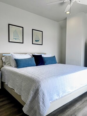 Una cama bien hecha con colcha blanca y almohadas azules, un ventilador de techo y dos cuadros en la pared.