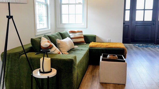 A living room with a green sectional sofa, a floor lamp, a side table with a candle, and a wooden bench. - Lux Lakeview Experience Sleep 8 (Hopatcong)