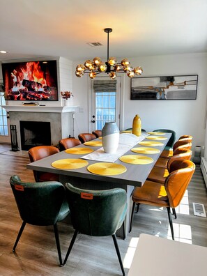 A dining area with a long table, multiple chairs, a television displaying a fireplace, and a modern ceiling light fixture. - Lux Lakeview Experience Sleep 8 (Hopatcong)