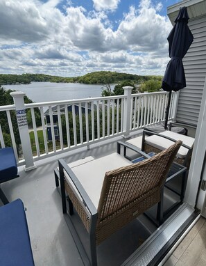 Een balkon met een tafel en stoelen, uitkijkend op een meer met zicht op bomen en huizen.