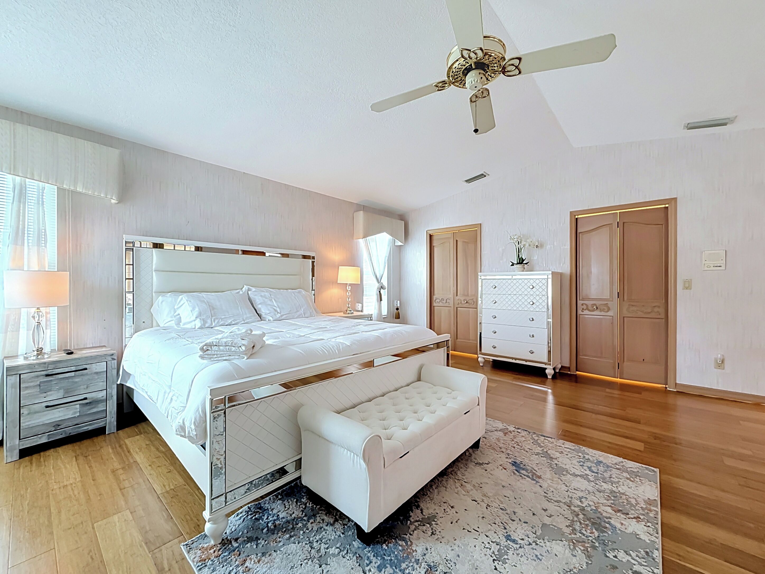 Une chambre à coucher comprenant un lit, des tables de chevet, une commode, un banc, une commode à tiroirs, une armoire et un ventilateur de plafond.
