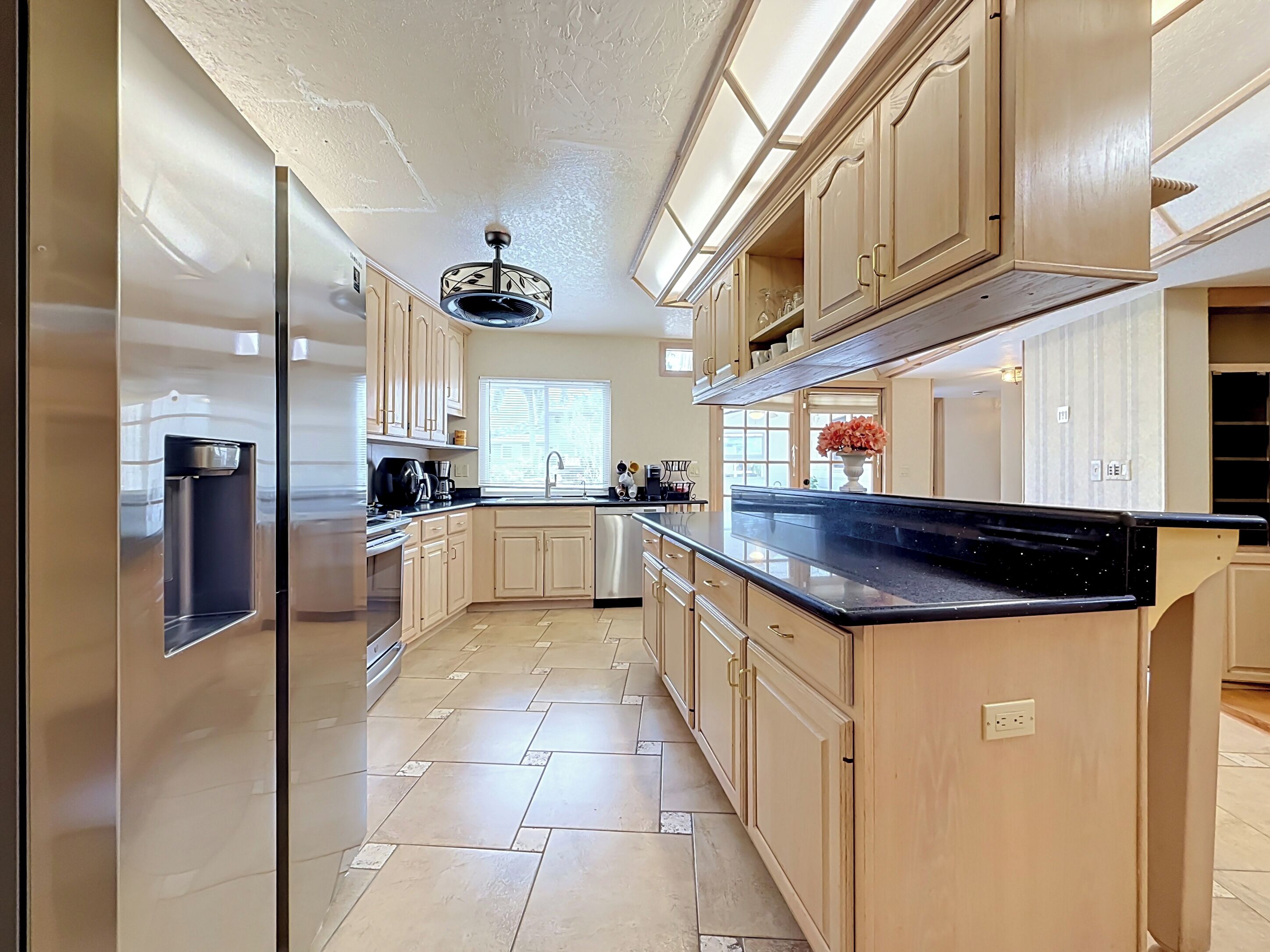 A modern kitchen with light wood cabinets, a black countertop, and a stainless steel refrigerator.