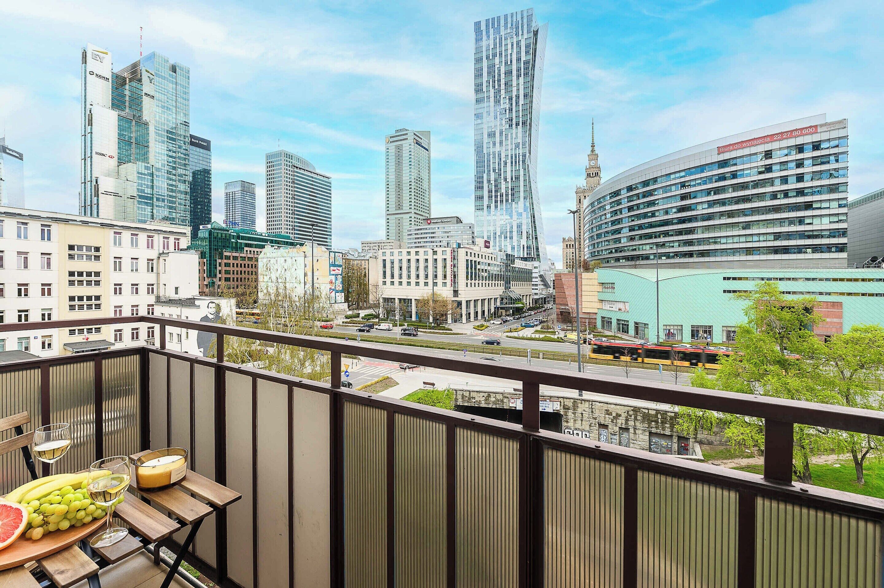 Ein Balkon mit Tisch für zwei Personen, Blick auf eine Skyline moderner Gebäude und einen klaren Himmel.