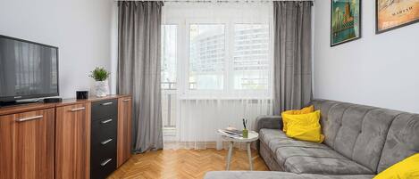 Una sala de estar moderna con un sofá gris, un televisor sobre un mueble de madera y una ventana con cortinas.