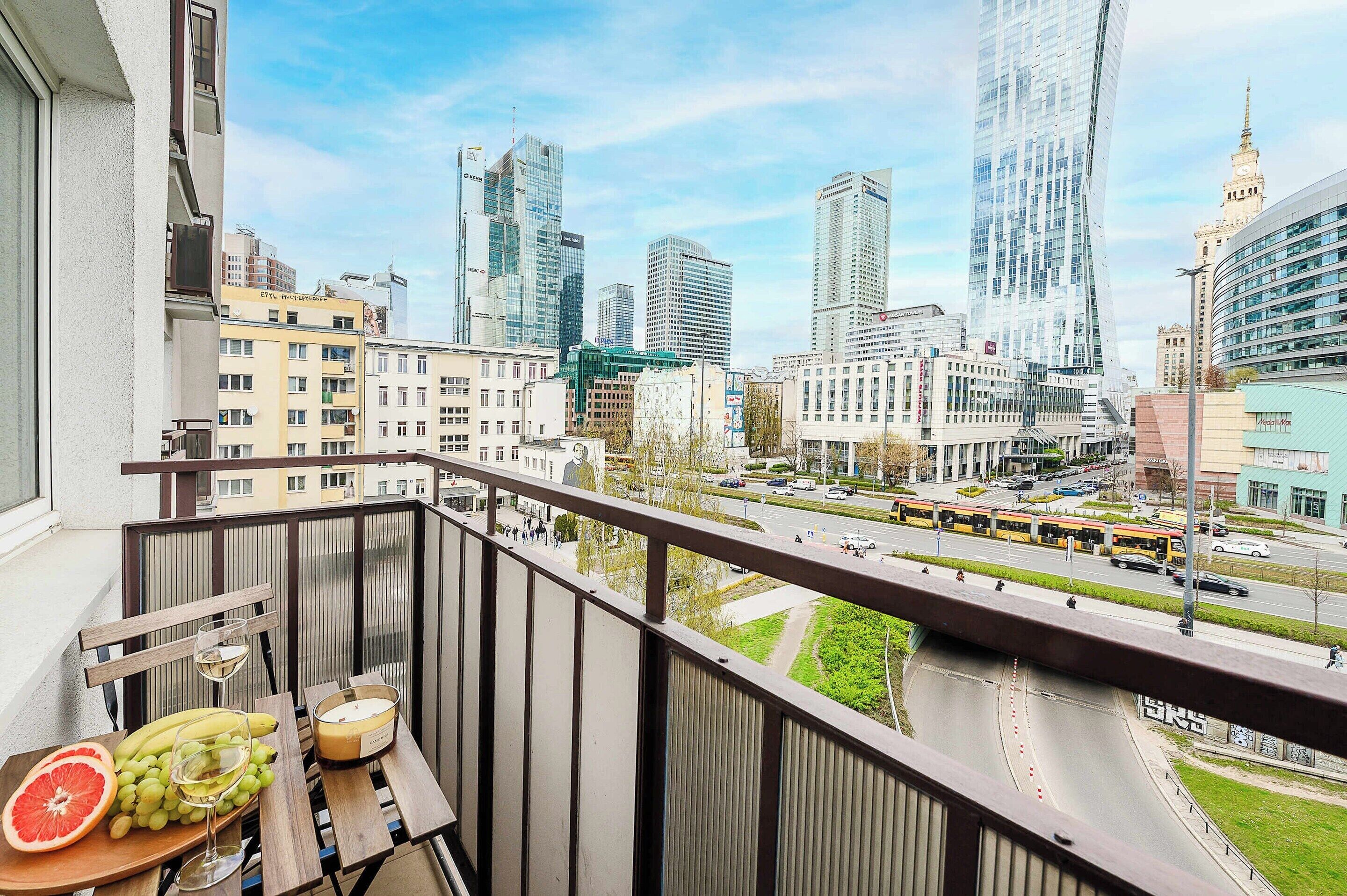 A balcony with a table set for dining, overlooking a cityscape with modern buildings and a busy street.