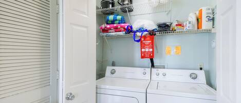 Une buanderie équipée de deux machines à laver à chargement frontal blanches, d’une étagère contenant divers objets, dont une machine à café, et d’une couverture anti-feu rouge accrochée au mur.