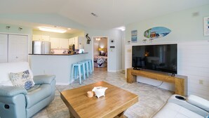 A living room with a sofa, a coffee table, a TV, and a kitchen in the background. - Pool & Hot Tub Access! Family Condo, 5 Mi to Beach (Gulf Shores)