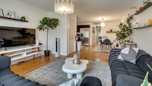 A modern living room with a flat-screen TV, a sofa, a coffee table, and a dining area in the background. - Easy NYC Commute: Furnished Jersey City Home (Jersey City)