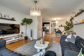 A modern living room with a flat-screen TV, a sofa, a coffee table, and a dining area in the background. - Easy NYC Commute: Furnished Jersey City Home (Jersey City)