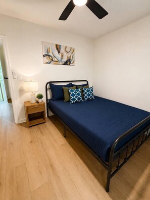 A single bed with a dark blue bedspread and patterned pillows, a bedside table with a lamp, and a ceiling fan. - The Sumerville's Carriage House (Summerville)