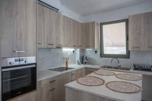 A modern kitchen with wooden cabinets, a built-in oven, a coffee machine, and a large window. - Cretan View | House with Valley & Mountain Views in Sitia, Crete (18' to Sitia) (Agios Georgios)