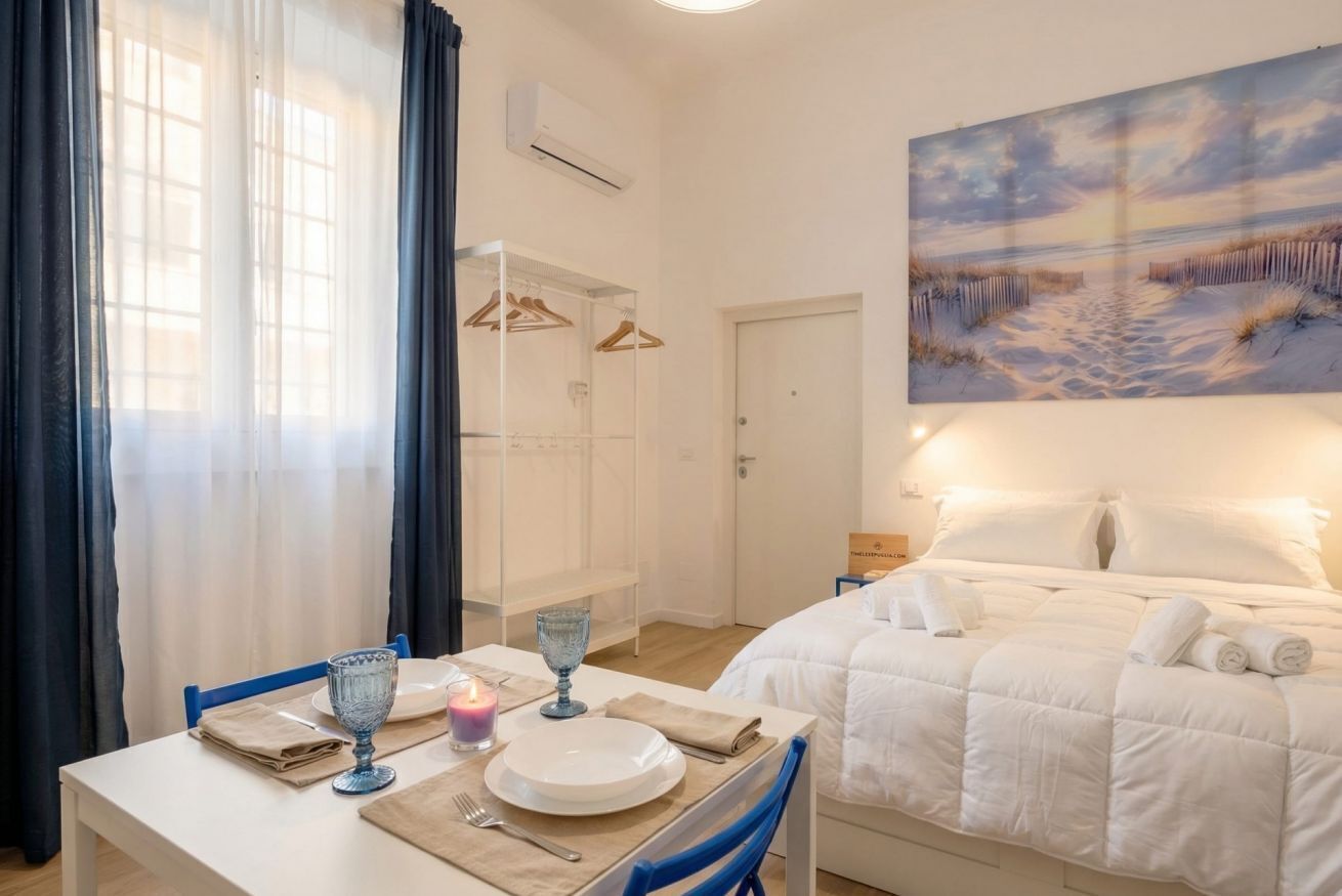 A hotel room with a bed, a dining table with a candle, and a painting of a beach scene.