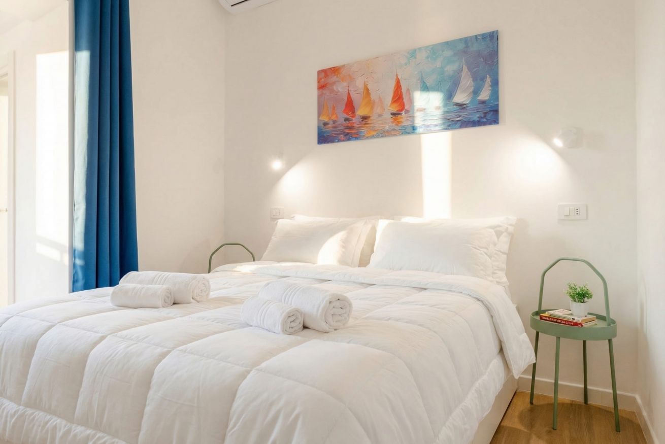 Uma cama bem arrumada com roupa de cama branca e toalhas enroladas, um quadro de veleiros na parede e uma pequena mesa de apoio verde com uma planta.