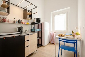 Eine kompakte Küche mit integrierter Kaffeemaschine, Wasserkocher und Kühlschrank. Es gibt eine Essecke mit Tisch und Stühlen, und ein Fenster sorgt für natürliches Licht.