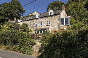 Ein Steinhaus mit vielen Fenstern, einem sichtbaren Schornstein und einem Garten mit dichter Bepflanzung.