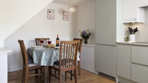 A dining area with a table set for two, wooden chairs, a bottle of wine, and a vase with flowers. - Claire's Cottage -  beautiful dog friendly cottage (Brimscombe)