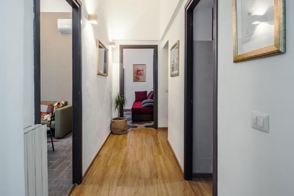 Un pasillo con piso de madera, un radiador y una puerta que da a una habitación con un sofá rojo y una planta.