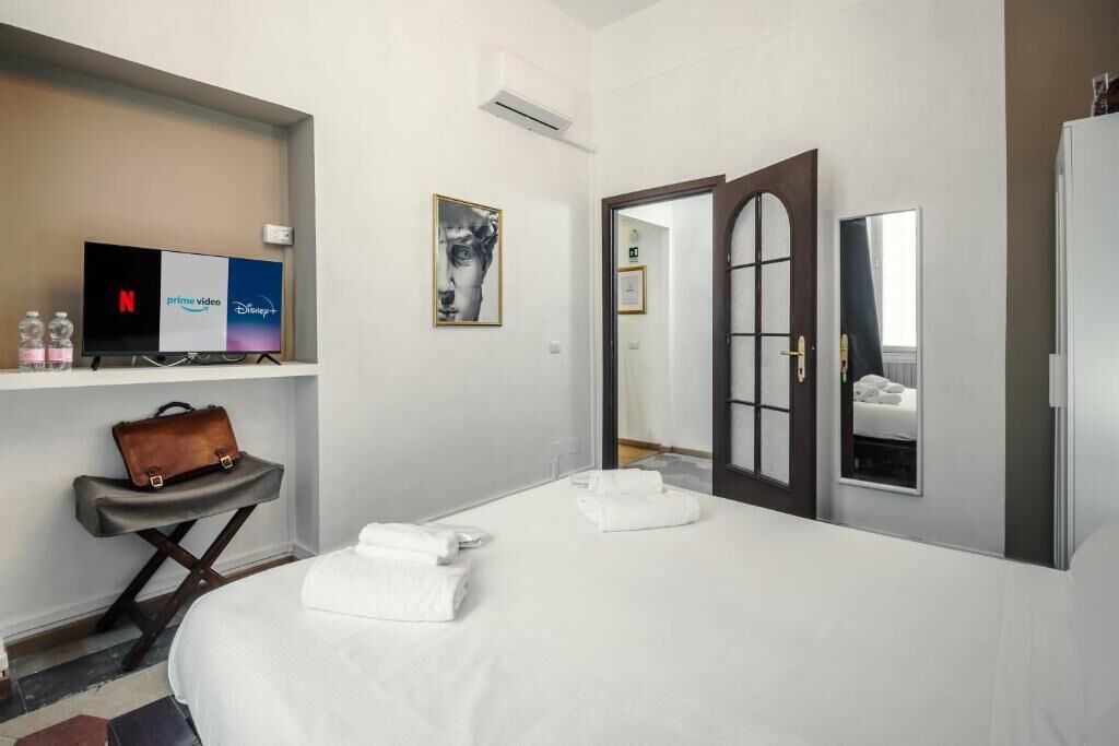 A hotel room with a bed, a desk with a laptop, a television displaying streaming services, and a door leading to another room.