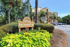 A wooden sign with 'OCEAN WALK ENTRANCE' and an arrow pointing right, surrounded by green foliage and a parking area with cars. - Ocean Oriented Oasis!  Indoor Heated Pool!  Pickleball! Hot Tub! (Hilton Head)