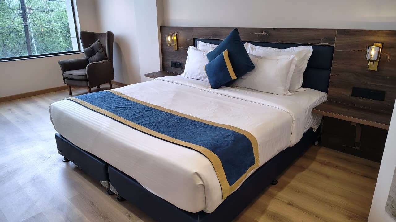 A hotel room with a large bed, a nightstand, a chair, and a window with a view of trees.