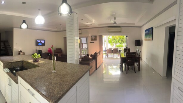 Una cocina moderna con armarios blancos, encimeras de granito y una isla central. Se observa un área de comedor con una mesa y sillas al fondo.