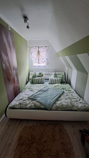 Ein Schlafzimmer mit einem Bett, einem Fenster mit Vorhängen, einem Heizkörper und einer Wand mit strukturierter Oberfläche.