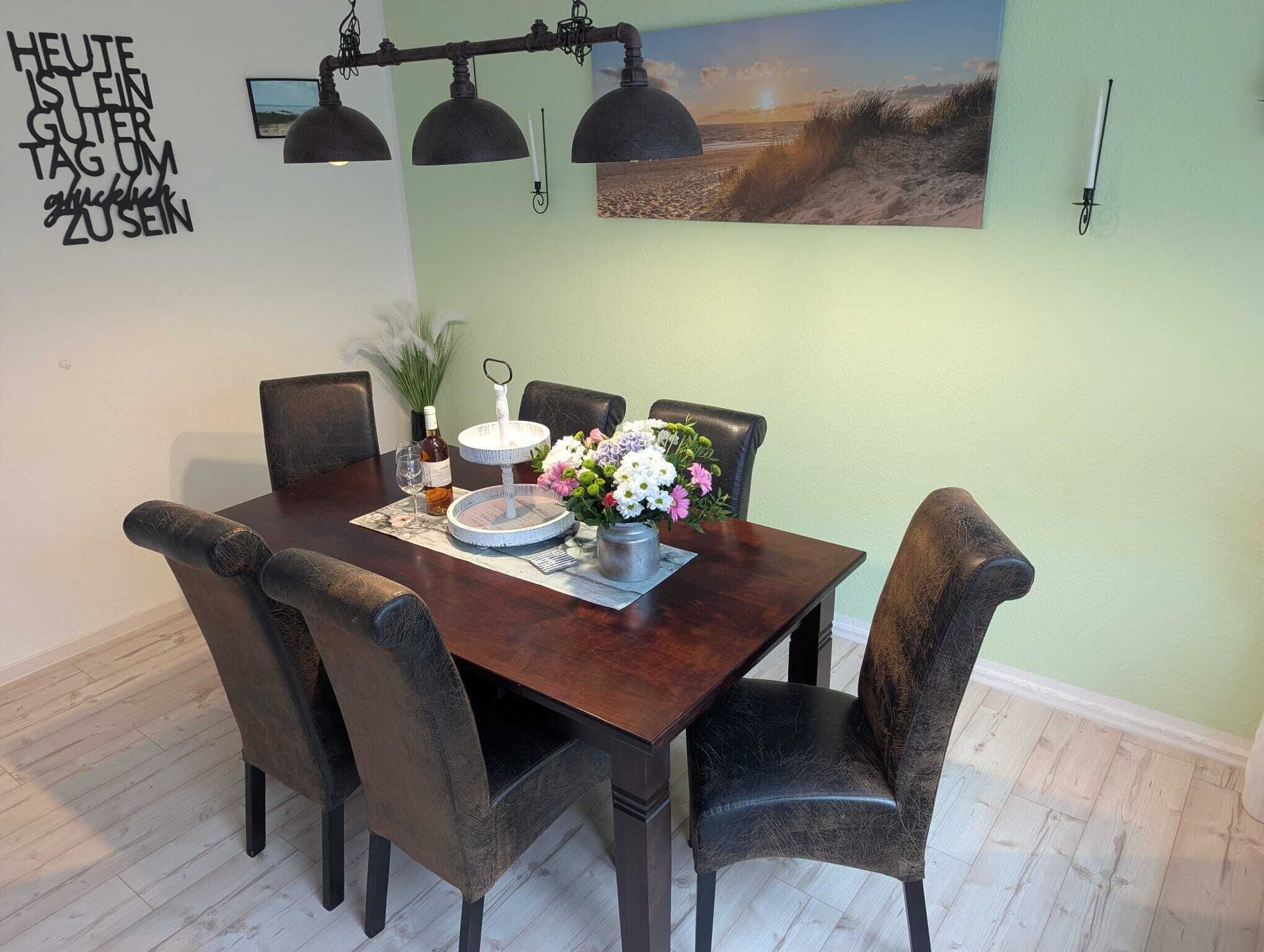 Uma sala de jantar com mesa de madeira, cadeiras escuras, um vaso de flores, uma garrafa de vinho e uma vela.