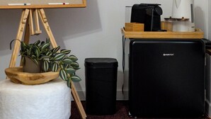 Um quarto com um cavalete de madeira, um banquinho pequeno com uma planta, uma lixeira preta, um alto-falante preto e uma cafeteira em uma mesa de madeira.