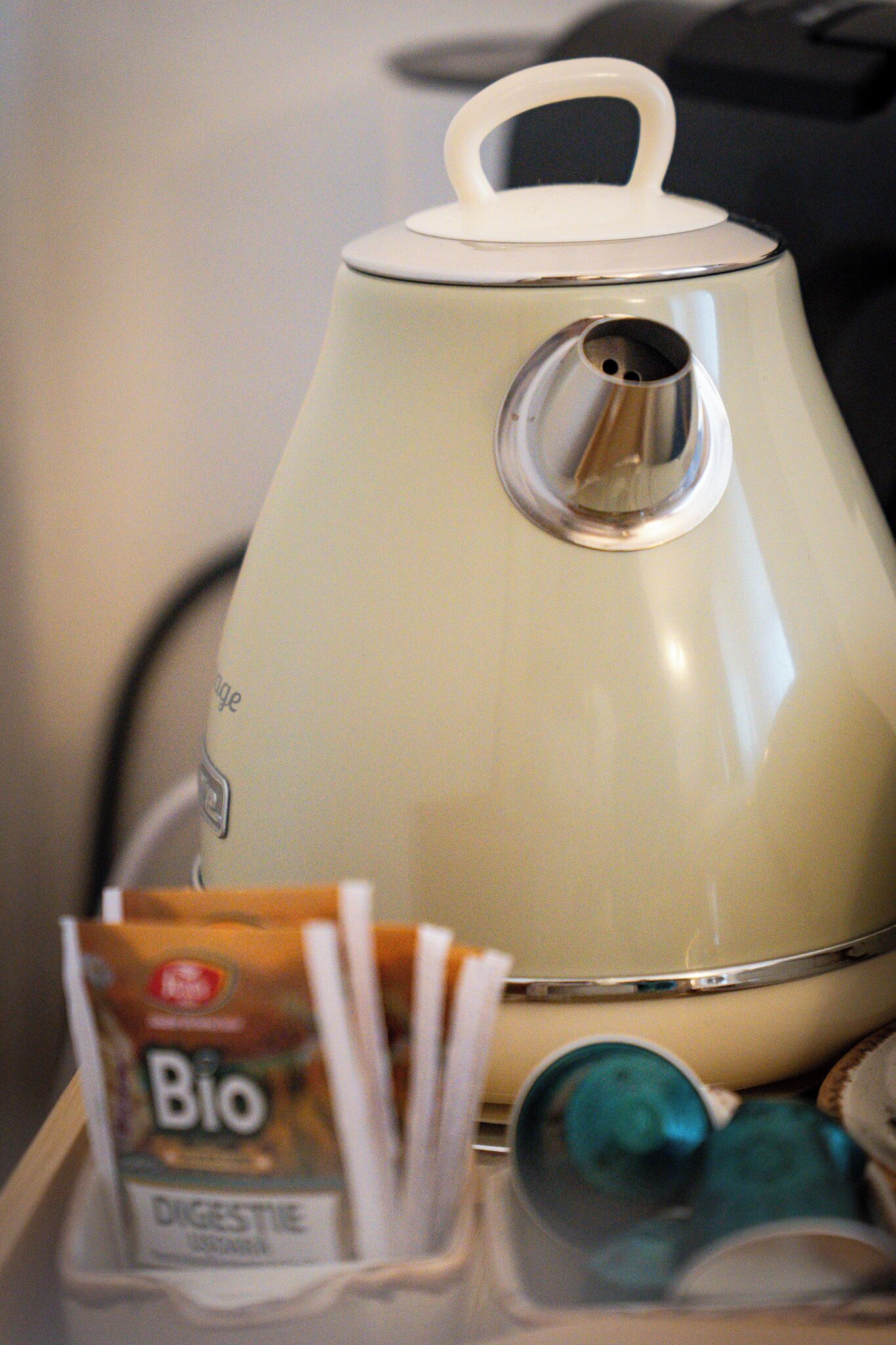 Una cafetera de estilo vintage con un asa blanco y un pico, sobre una mesa con una caja de cápsulas de café etiquetada 'Bio Digestive' y un objeto azul verdoso a su lado.
