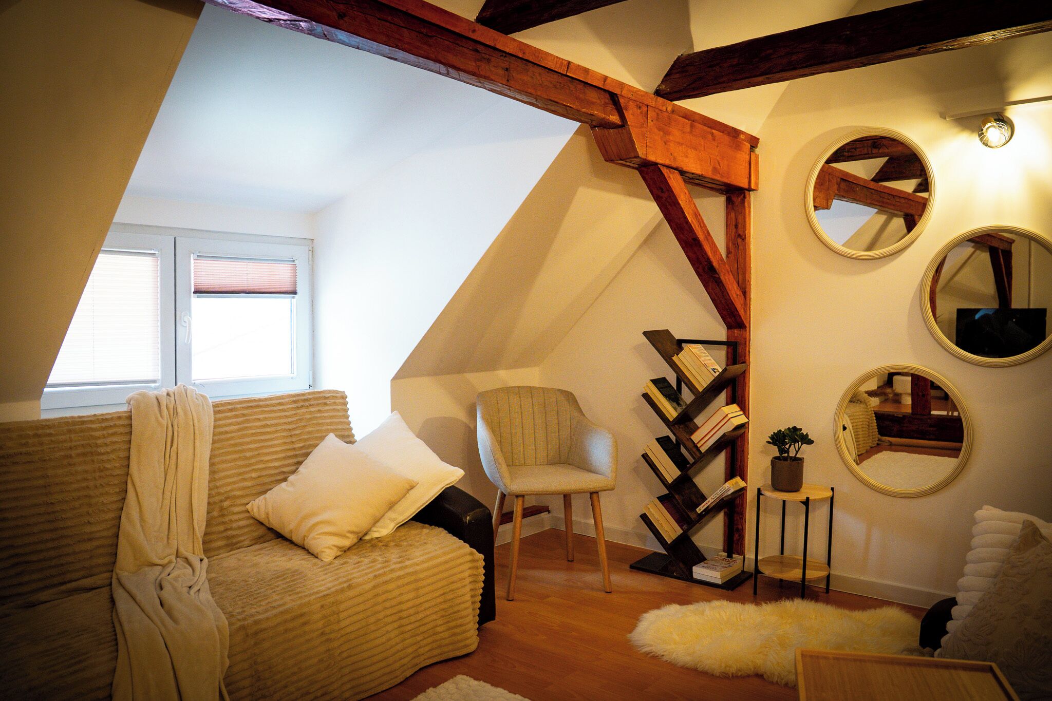 A cozy attic room with a sloped ceiling, a cushioned sofa, a chair, a small table with a plant, and a bookshelf.
